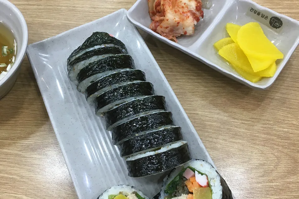 Overhead view of a sliced gimbap roll on a white plate with kimchi and yellow pickled radish on the side, on a light wooden table.