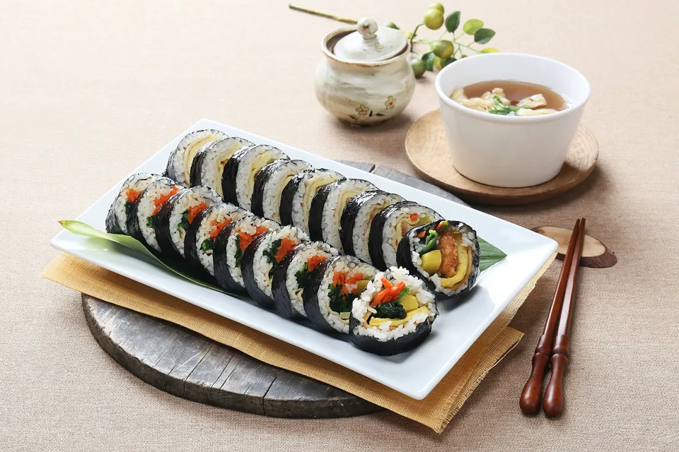Neatly plated gimbap slices on a white rectangular dish on a wooden board, with a bowl of soup, chopsticks and a beige linen backdrop.